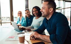 Two men and two women sitting in a meeting, one typing on a laptop.